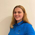 ALDI SÜD Azubine im blauen Polohemd steht vor einer weißen Wand und lächelt freundlich in die Kamera