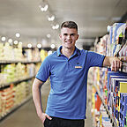  ALDI SÜD Auszubildender stützt seinen rechten Arm auf ein Regal in der Filiale und blickt freundlich in die Kamera