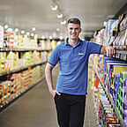  ALDI SÜD Auszubildender stützt seinen rechten Arm auf ein Regal in der Filiale und blickt freundlich in die Kamera