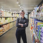 ALDI SÜD Mitarbeiterin steht mit verschränkten Armen im Gang einer Filiale und lächelt in die Kamera