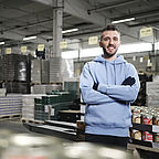 ALDI SÜD Mitarbeiter steht mit verschränkten Armen in der Logistikhalle und lächelt freundlich in die Kamera