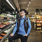 ALDI SÜD Auszubildender steht am Kühlregal, ein Bein auf dem Regal, die Hand in der Tasche und lächelt in die Kamera