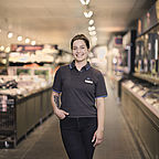 ALDI SÜD Mitarbeiterin steht mit einer Hand in der Tasche im Gang einer Filiale und lächelt freundlich in die Kamera