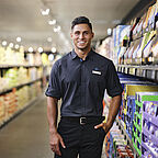 ALDI SÜD Filialleiter steht im Gang der Filiale neben einem Regal, die rechte Hand in der Tasche und lächelt in die Kamera