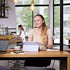 Junge ALDI SÜD Mitarbeiterin sitzt mit einer Tasse Kaffee, Notizbuch und Tablet an einem Tisch in der Kantine und lächelt